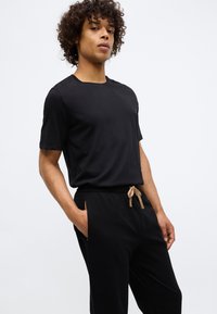 Crna pamučna majica s okruglim izrezom u kombinaciji s crnim hlačama koje imaju bež vezicu i bočne džepove. Glatka tekstura.