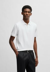 White short-sleeve polo shirt in textured fabric with a zip closure, worn with black trousers and a belt featuring a silver buckle.