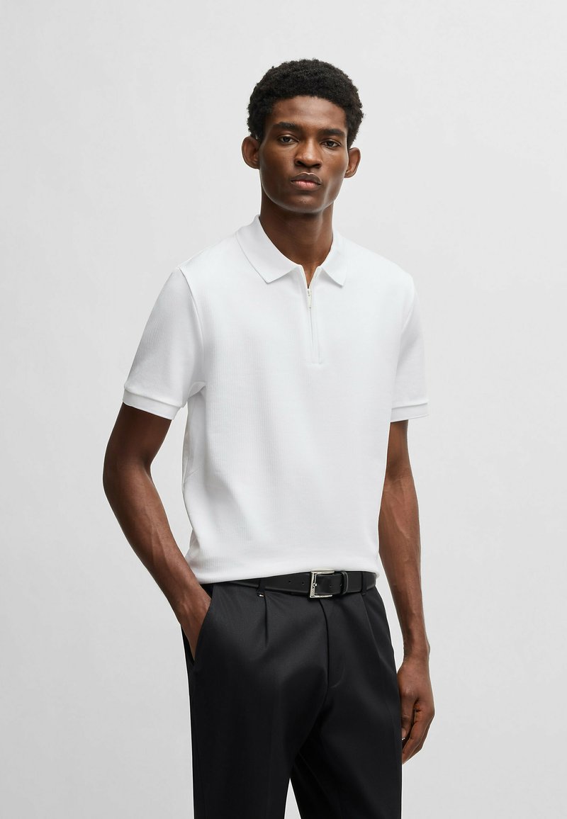 White short-sleeve polo shirt in textured fabric with a zip closure, worn with black trousers and a belt featuring a silver buckle.
