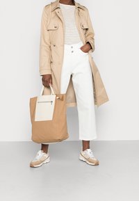 Cappotto trench beige sopra una camicia a righe, abbinato a pantaloni bianchi a gamba larga e sneaker beige, mentre tiene una borsa a tracolla color sabbia con un pannello crema e zip.