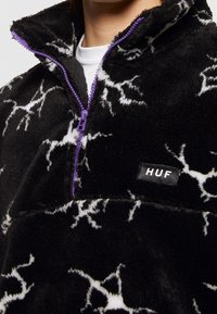 Pull polaire noir avec motifs d’éclairs blancs, fermeture éclair violette et écusson avec le logo HUF sur la poitrine, porté sur une chemise blanche.