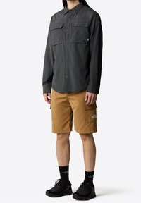 Camicia grigia con bottoni e due tasche sul petto, abbinata a pantaloni cargo beige con tasche laterali. Sneakers nere e calzini completano l'outfit.