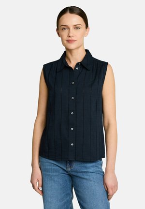 Femme aux cheveux foncés portant une chemise noire sans manches boutonnée et un jean bleu, debout devant un fond clair et uni.