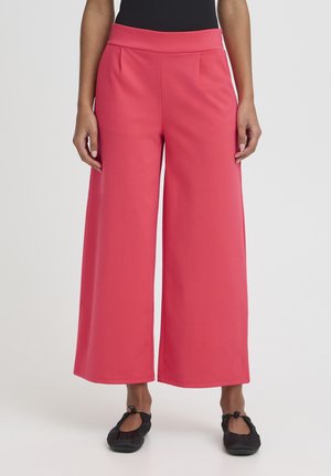 Mujer con pantalones coral de talle alto y pierna ancha, llevando bailarinas negras, posando frente a un fondo blanco liso.