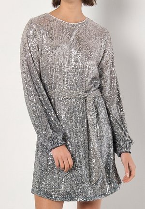 Kobieta ubrana w srebrną, cekinową suknię z długim rękawem i paskiem wiązanym w pasie, stojąca na tle jednolitego jasnego tła.