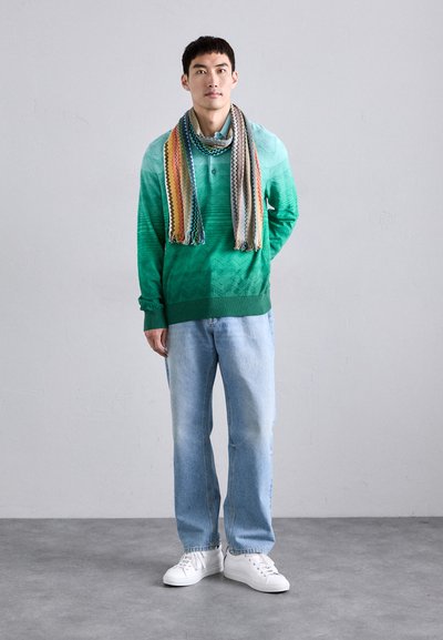 Sweater em degradê verde com padrão texturizado, combinado com jeans azuis claros e ténis brancos. Cachecol estampado multicolorido drapeado em torno do pescoço.