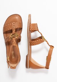 Sandales en cuir brun avec des sangles tressées, accent en logo doré, talon plat et semelle cousue. Design ouvert pour la respirabilité.