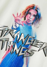 Hvid t-shirt med et grafisk print af en pige med bølget hår, der rækker ud, omgivet af "STRANGER THINGS" i fed, sort skrift.