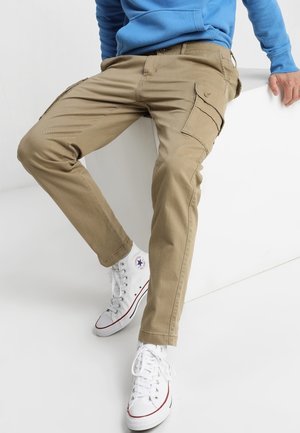 Personne assise sur une surface blanche, portant un pantalon cargo beige, des baskets montantes blanches et un sweat bleu, jambes croisées aux chevilles.