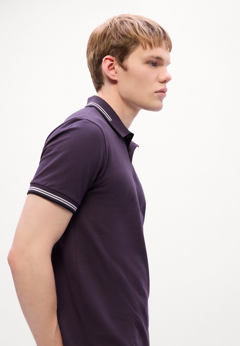 Jeune homme aux cheveux blonds courts portant un polo violet foncé avec des rayures blanches, tourné vers la droite sur un fond blanc uni.