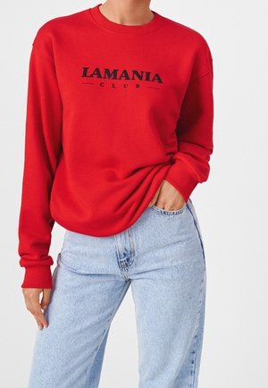Person trägt roten Pullover mit langen Ärmeln und der Aufschrift "LAMANIA CLUB" sowie hellblaue Jeans mit hoher Taille, Hand in der Tasche.