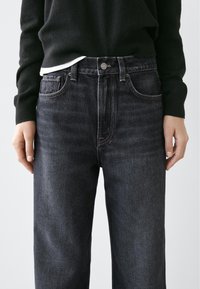 Tmavě modré džíny z pevného denimu s vysokým pasem, zapínáním na knoflíky, tradičním pětikapsovým designem a jemným vyblednutím. Doplněné černým svetrem.