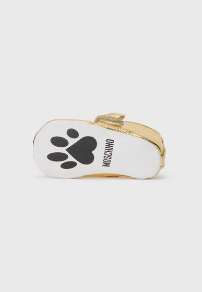 Zapato dorado con suela blanca que presenta una huella de pata negra. Incluye una correa en la parte superior y una textura suave para un detalle adicional.