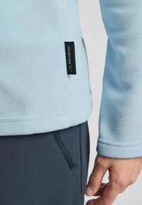 Pullover in pile blu chiaro con orlo arrotondato, cuciture piatte e etichetta Polartec nera e verde sul lato, abbinato a pantaloni blu scuro.