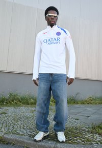 Hvid langærmet sportstrøje med blå accenter og "QATAR AIRWAYS" logo, parret med løse blå jeans og hvide sneakers, udendørs.