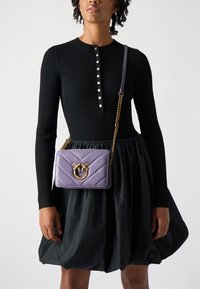 Bolsa de mão acolchoada roxa com ferragens douradas e uma alça de corrente, usada com um vestido preto canelado com detalhes em botão e uma saia ampla.