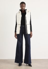 Pufferväst i off-white med huva och justerbara snören, kombinerad med mörkblå vida jeans och spetsiga svarta klackar.