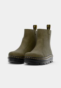 Bottines vert olive en cuir texturé avec une semelle noire épaisse et des coutures jaunes. Comprend des tirettes pour un enfilage facile.