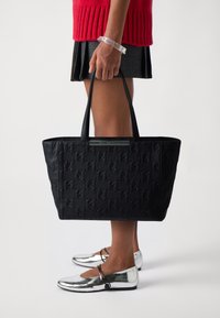 Žena v stříbrných balerínách, s černou kostkovanou sukní, červeným svetrem a průhledným náramkem, držící velkou černou tašku přes rameno s vyraženým vzorem.