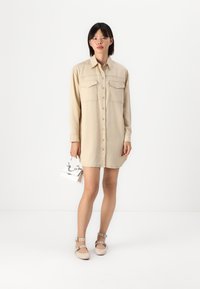 Abito camicia beige con maniche lunghe, due tasche sul petto, chiusura frontale con bottoni e una vestibilità ampia, abbinato a scarpe beige con cinturini e una piccola borsa bianca.