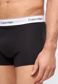 Miehen vyötärö ja yläreisi mustissa Calvin Klein -bokserialushousuissa, joissa valkoinen brändätty vyötärönauha, oikea käsi rentona sivulla.