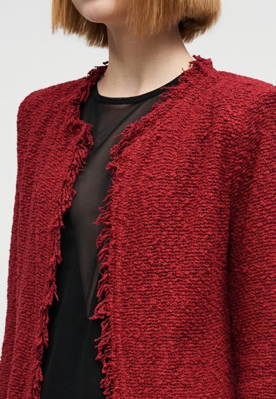 Κόκκινο υφασμάτινο μπουφάν με ξεφτισμένα άκρα, που διαθέτει στρογγυλή λαιμόκοψη. Φορεμένο πάνω από ένα μαύρο διάφανο τοπ, αναδεικνύοντας τα αντίθετα υλικά.