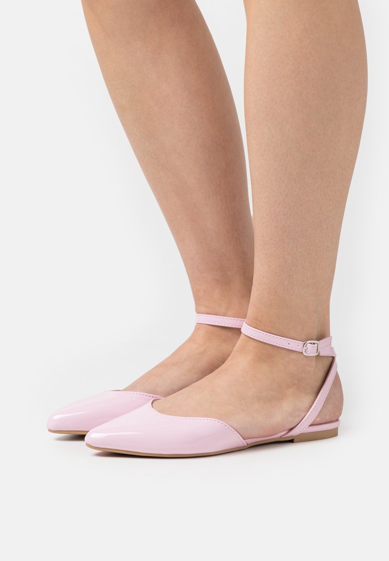 Ballerines rose clair à bout pointu avec des lanières à la cheville, portées pieds nus sur un fond blanc.