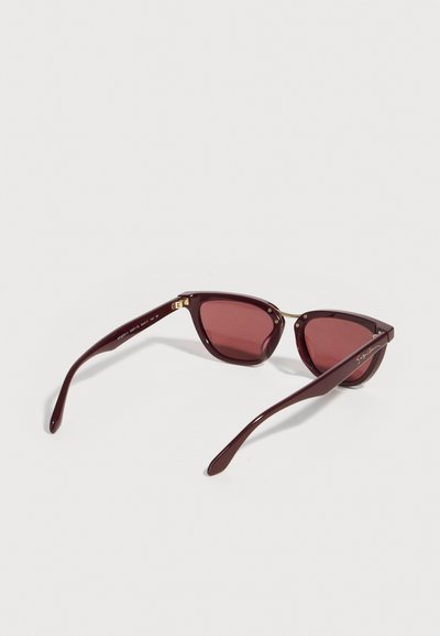 Σκούρα κόκκινα ορθογώνια γυαλιά ηλίου με χρωματισμένους φακούς και λεπτές χρυσές λεπτομέρειες, εμφανίζονται να ακουμπούν σε λευκή επιφάνεια από πλάγια οπίσθια όψη.