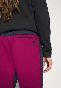 Vinröda träningsbyxor med marinblå accenter, med en ficka med dragkedja och en liten grön tryckt logotyp på baksidan. Närbild av midjebandet och hand.