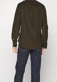 Mann, der ein dunkelgrünes Langarmhemd und blaue Denim-Jeans trägt, von hinten vor einem einfarbigen weißen Hintergrund gezeigt.