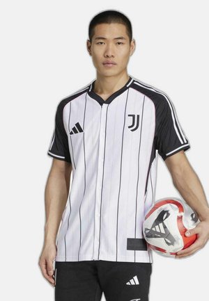 Camiseta de rayas blancas y negras con cierre de botones, que presenta un logo en el pecho y un panel texturizado negro. El jugador sostiene un balón de fútbol rojo y blanco.