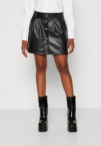 Jupe en simili cuir noir avec une forme évasée, taille élastique, fermeture à boutons sur le devant et finition brillante. Associée à des bottines noires brillantes.