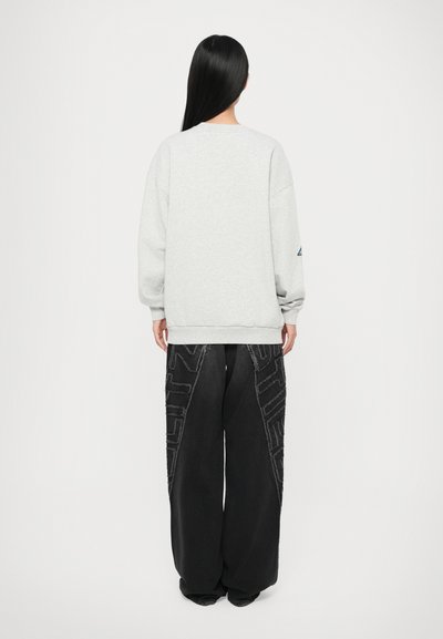Sweatshirt oversized grigio chiaro con spalle cadenti e polsini a coste, abbinato a pantaloni larghi neri con motivo geometrico testurizzato.