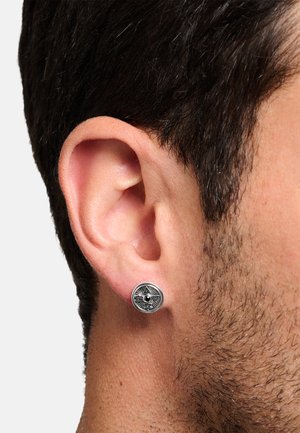 Ohrringe aus silberfarbenem Metall mit kreisförmigem Design, das geometrische Muster und einen zentralen schwarzen Stein aufweist. Angenommen an einem Ohr eines Modells.