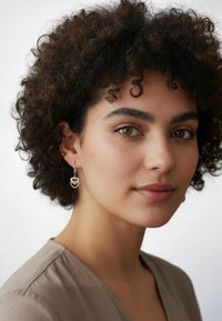 Orecchini a cerchio in argento con charm a forma di cuore caratterizzati da un design a strati. Il modello ha i capelli ricci e indossa una maglietta beige.