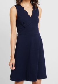 Marinblå ärmlös klänning med en scalloped halsringning, åtsittande bodice och en utsvängd A-linje kjol, gjord av mjukt, stretchigt material.