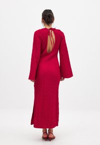 Red, textured long-sleeve dress with a back cutout and tie detail. Features a straight hem and side slit. Worn with pink heeled sandals.