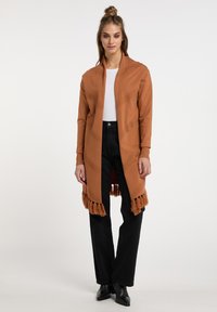 Lange, roestkleurige cardigan met een gestructureerd zigzagpatroon en franjes aan de onderkant. Draagt over een witte top en gecombineerd met zwarte broek en laarzen.
