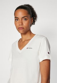 T-shirt bianco a V in cotone morbido, con un piccolo logo ricamato "Champion" in rosso e nero sul petto e sulla manica.