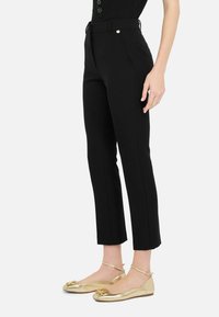 Schwarze maßgeschneiderte Hose mit schmaler Passform, ausgestattet mit einem Gürtel, Fronttaschen und einer glatten Textur. Kombiniert mit glänzenden goldenen Ballettschuhen.