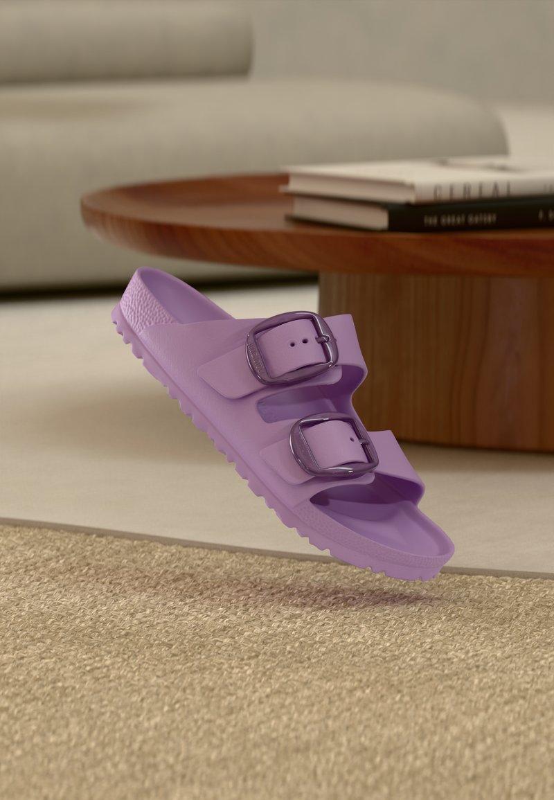 Lavendel dobbeltspandlet slip-in sandal svæver over en struktureret beige måtte, med rundt træ- sofabord og sofa i baggrunden.