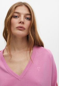 Rosa V-Ausschnitt-Pullover aus glattem Stoff, mit gerippten Kanten und einem kleinen gestickten Herzdetail auf der linken Seite.
