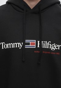 Черна суитчър с лого Tommy Hilfiger в бяло и правоъгълна емблема на флаг в червено, синьо и бяло, и текст "Authentic Originals since 1985" в червено.