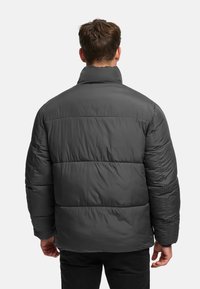 Man van achteren gezien, draagt een donkergrijze donzen jas en zwarte broek, staand tegen een effen lichte achtergrond.