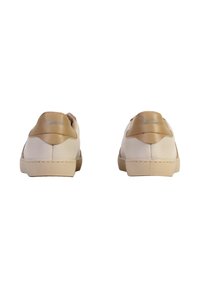 Un par de zapatillas blancas de caña baja con acentos en el talón beige, mostradas desde la parte de atrás sobre un fondo blanco.