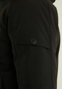 Gros plan sur un tissu noir montrant une poche à rabat avec un bouton rond noir sur le côté d'un vêtement.