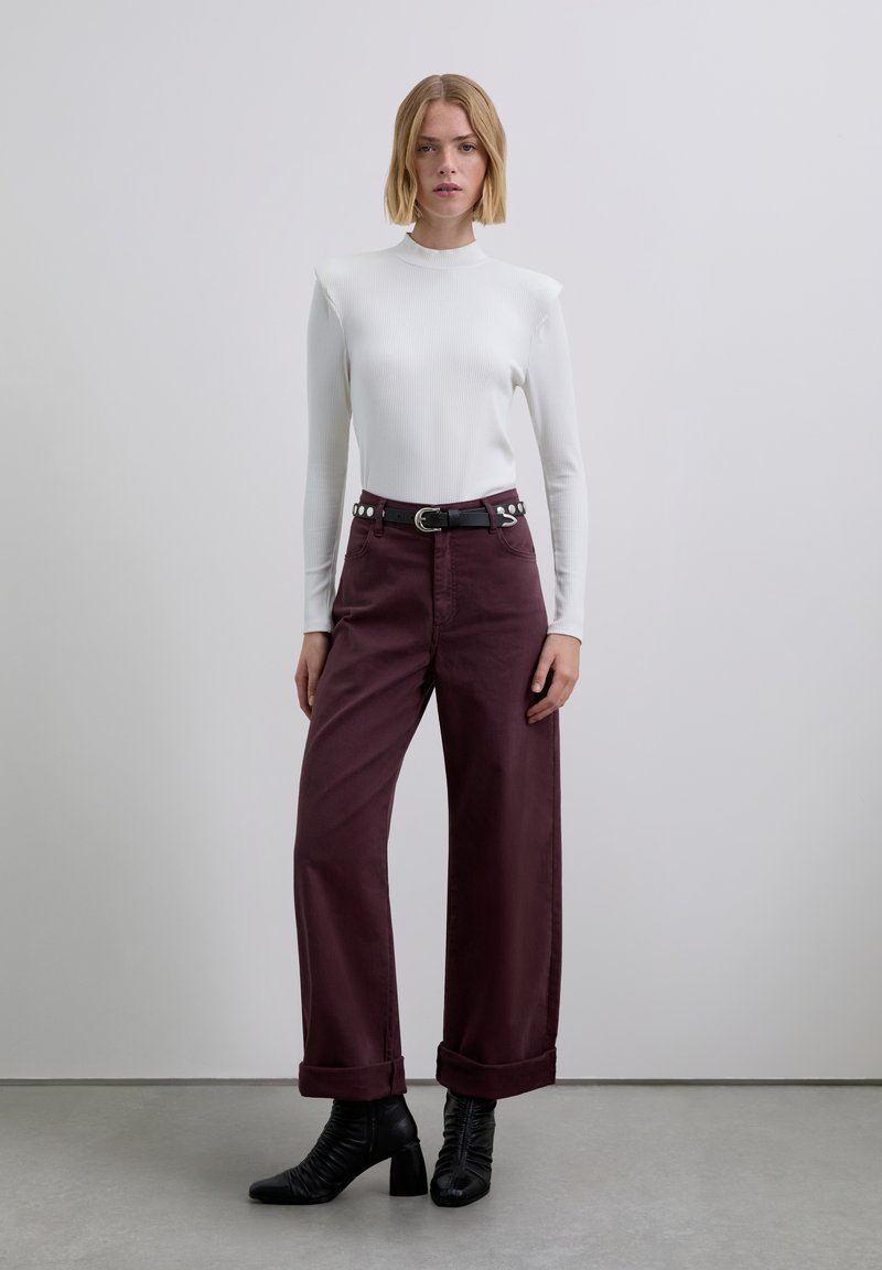 Witte geribde top met lange mouwen gecombineerd met wijde, bordeauxrode broek met opgerolde pijpen en een zwarte riem met zilveren details. Zwarte enkellaarzen met hak.