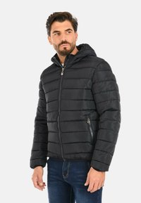 Uomo che indossa un giubbotto a vento trapuntato nero con cappuccio, dotato di zip frontale e tasche con zip, abbinato a jeans blu scuro, che guarda di lato.