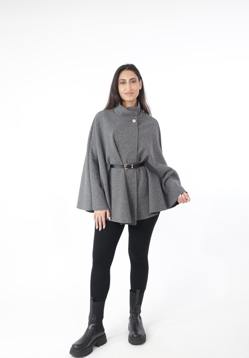 Mantella in misto lana grigia con collo alto, caratterizzata da un bottone dorato e una cintura nera, abbinata a leggings neri e stivali neri robusti.