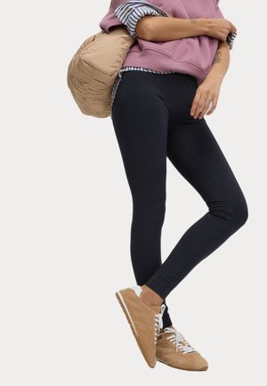 Marineblauwe leggings met een gladde textuur, gecombineerd met een lichtroze oversized sweatshirt en bruine sneakers, terwijl een beige puffer tas wordt vastgehouden.